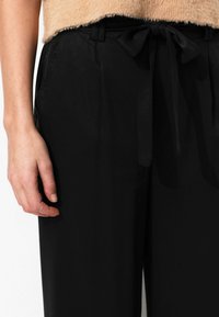 Pantalons noirs taille haute avec une ceinture en tissu nouée. texture douce, poches latérales et coupe décontractée. Gros plan d'une main sur la taille.