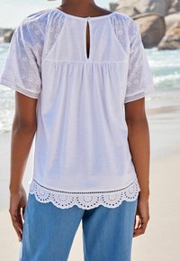 Weißes Oberteil mit bestickten Netzärmel, Schlüsselloch-Rücken, gerafftem Brustbereich und gewelltem Saum. Kombiniert mit hellblauer Denim.