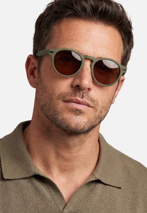 Uomo che indossa occhiali da sole rotondi verde oliva e una camicia verde oliva con colletto abbinata, che guarda leggermente di lato.