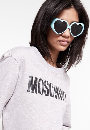 Donna con capelli scuri fino alle spalle che indossa occhiali da sole azzurri a forma di cuore e una felpa grigio chiaro con "MOSCHINO" stampato in nero.