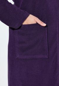 Ruka vložená do přední kapsy dlouhého, tmavě purpurového fleecového oděvu s viditelným zipem a texturovanou tkaninou.