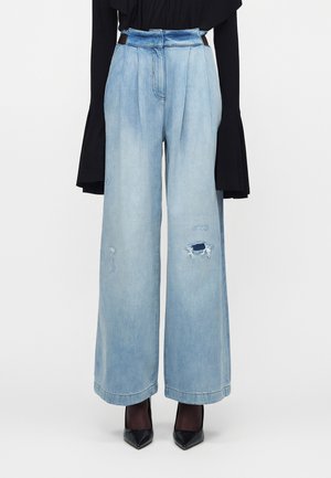 Φαρδιά τζιν παντελόνια ανοιχτού μπλε χρώματος με φθαρμένες περιοχές, φορεμένα με μαύρες ψηλοτάκουνες γόβες και μαύρη μπλούζα με μακριά μανίκια.