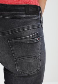 Jeans de mezclilla negra con un diseño de bolsillo trasero curvado, que presenta detalles de costura sutiles y una etiqueta de cuero negro con marca en la cinturilla.