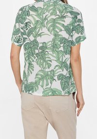 Camicia a maniche corte con sfondo bianco e stampa di foglie tropicali verdi. Tessuto fluido, vestibilità morbida e scollo rotondo, abbinata a pantaloni beige.