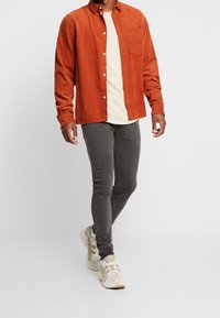 Camisa de pana de color óxido con bolsillo frontal, llevada sobre una camiseta crema. Combinada con jeans gris ajustados y zapatillas deportivas ligeras.