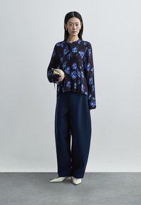 Mulher em pé a usar uma blusa preta com padrões florais azuis, calças largas azul-marinho, sapatos brancos pontiagudos e a segurar uma pequena carteira bege.