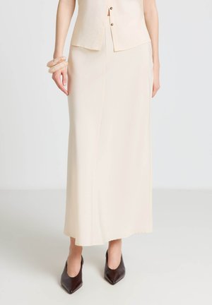 Femme portant une longue jupe crème avec un gilet assorti et des chaussures à bout pointu foncé, debout devant un fond uni.