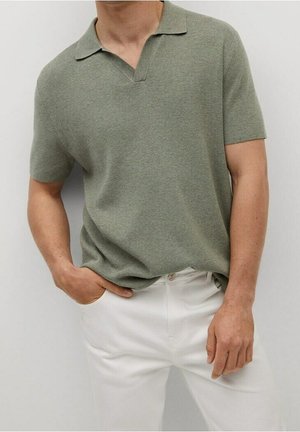 Homme portant un polo en maille vert olive à manches courtes avec col ouvert, rentré dans un pantalon blanc, debout avec une main dans la poche.