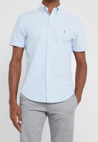 Ljusblå kortärmad skjorta med knappar i bomull, med klassisk krage, prydd med en liten logotyp på vänster bröst och en rak fåll.