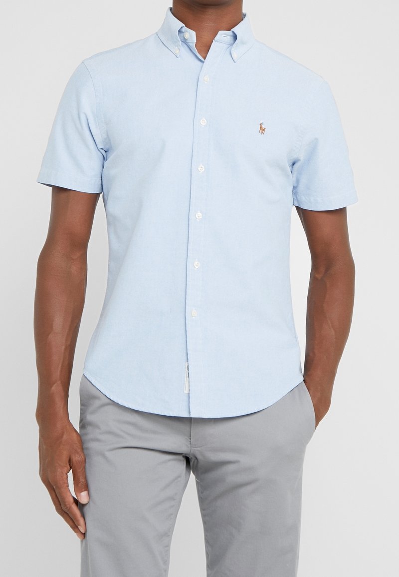 Ljusblå kortärmad skjorta med knappar i bomull, med klassisk krage, prydd med en liten logotyp på vänster bröst och en rak fåll.