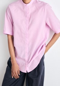 Różowa koszula z krótkim rękawem z kołnierzem typu mandarynka, z przodem zapinanym na guziki i subtelną fakturą; w połączeniu z ciemnymi spodniami.