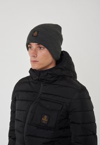 Giubbotto puffo nero con cuciture orizzontali e una tasca frontale con toppa del logo. Berretto lavorato a maglia grigio scuro con texture a coste.
