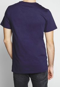 Mies, joka käyttää yksinkertaista tummanpunaista lyhythihaista t-paitaa ja mustia farkkuja, nähtynä selkäpuolelta vaaleaa taustaa vasten.