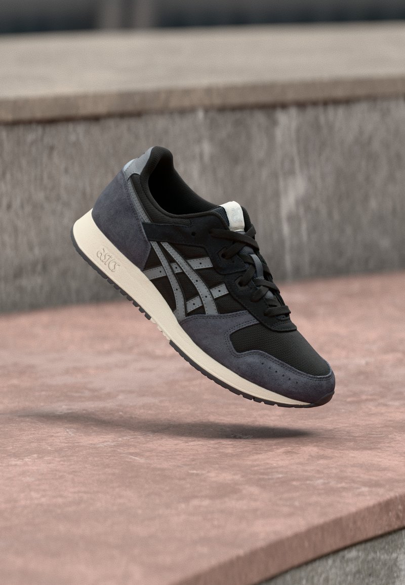 Svart och grå mocka och mesh sneaker med en vit mellansula, med en distinkt sidologga och texturerade accenter, svävande ovanför en yta.