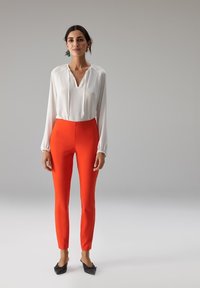 Blouse blanche à manches longues avec un col en V et des poignets froncés, associée à un pantalon orange ajusté. Des ballerines noires à bout pointu complètent la tenue.