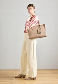 Beige leren tote bag met handvatten aan de bovenkant, rechthoekige vorm en een decoratief label; gedragen met een rood-wit gestreepte trui en crèmekleurige broek.