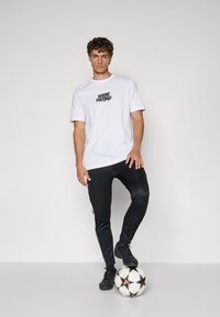 Hvid T-shirt med sort "Adidas Football" logo, parret med sorte joggingbukser med hvide side-striber. Sorte fodboldsko. Fodbold ved fødderne.