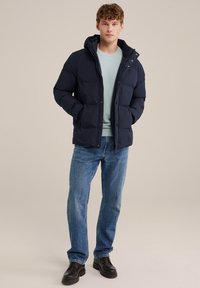 Navy puffer jacket with a hood, featuring a quilted texture and snap closures, paired with a light blue sweater and blue jeans, worn with black shoes.