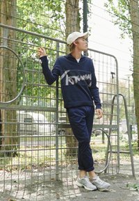 Granatowy sweter z jasnoszarym logo "Tommy", w połączeniu z granatowymi dresami i jasnoszarymi trampkami; osoba opiera się o metalowy płot.