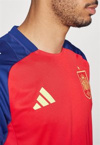 Červený a modrý sportovní dres, s krátkým rukávem, vyrobený z hladkého materiálu. Na přední straně se nachází žluté logo Adidas a státní znak Španělska.