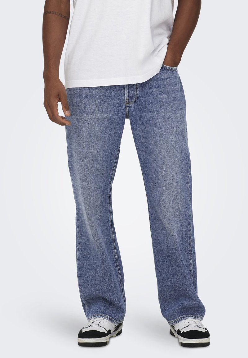 Blue denim jeans with a loose fit, featuring a classic five-pocket design, stitched seams, and subtle fading, paired with black and white sneakers.