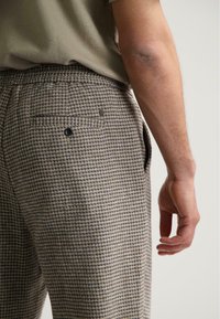 Beige en grijs geblokte broek gemaakt van een gestructureerde stof. Heeft een achterzak met een knop en wordt getoond met een gedeeltelijk zichtbare hand.