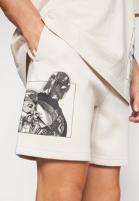 Calções bege claro feitos de tecido macio com uma estampa gráfica a preto e branco de uma personagem e uma criatura na perna esquerda.