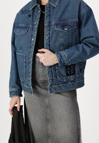 Chaqueta de mezclilla en azul oscuro con dos bolsillos en el pecho, botones en el frente y bordado floral en la manga. Llevada sobre una blusa negra y una falda gris.