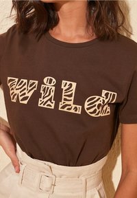 Brun bomullströja med ordet "WILD" i beige med ett zebramönster, korta ärmar och en avslappnad passform.