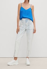 Helle blaue Jeans mit seitlichen Reißverschlüssen, kombiniert mit einem leuchtend blauen Tanktop. Das Model trägt weiße Sneakers und zeigt einen lässigen Look.