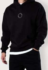 Czarna bluza z kapturem z przednią kieszenią typu kangurka i ściągaczami na mankietach. Na piersi znajduje się okrągłe białe logo. Materiał wydaje się miękki i trwały.