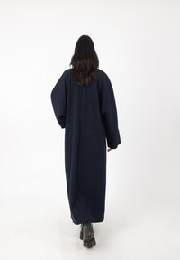 Abrigo azul marino de corte oversized con una silueta fluida, que cuenta con mangas anchas y una costura que recorre el centro de la espalda. Botines negros visibles.