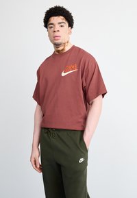 Bordo kokvilnas T-krekls ar īsām piedurknēm un balto Nike logotipu; apvienots ar olīvzaļiem sporta biksēm, kam ir neliels balts logotips.