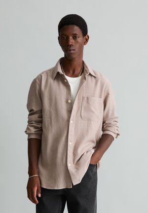Jeune homme à la peau foncée portant une chemise beige boutonnée sur un t-shirt blanc et un pantalon noir, debout avec une main dans la poche sur un fond gris.