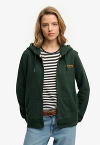 Sudadera verde con cremallera, capucha y bolsillos laterales, con tejido texturizado y un logo dorado "SuperDry" en el pecho, sobre una camiseta a rayas.