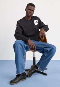 Svart hettegenser med hvit logo på brystet, sammen med løse blå denim jeans og svarte joggesko. Modell sittende på en tremodul.