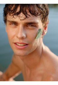 Jeune homme avec des cheveux courts et mouillés de couleur sombre et une feuille vert olive sur la joue. Peau lisse avec des reflets subtils, yeux clairs et expression neutre.