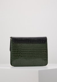 Suorakulmainen tummanvihreä ja musta krokotiilinahkaa jäljittelevä nahkalaukku, jossa on läppä, seisoo valkoisella pinnalla yksiväristä taustaa vasten.