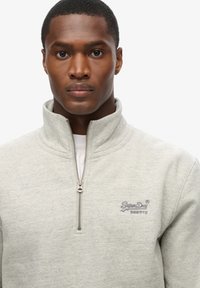 Sudadera de forro polar gris con cuello alto, cierre de cremallera en el cuarto frontal y logotipo bordado. La tela tiene una textura suave y un sutil patrón jaspeado.