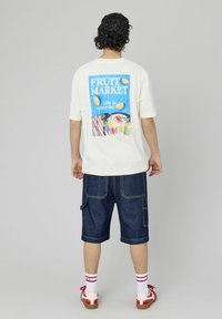 T-shirt en coton de couleur crème avec un graphique "FRUIT MARKET" en bleu, présentant des fruits colorés. Associé à un short en denim foncé et à des chaussettes rayées rouges.