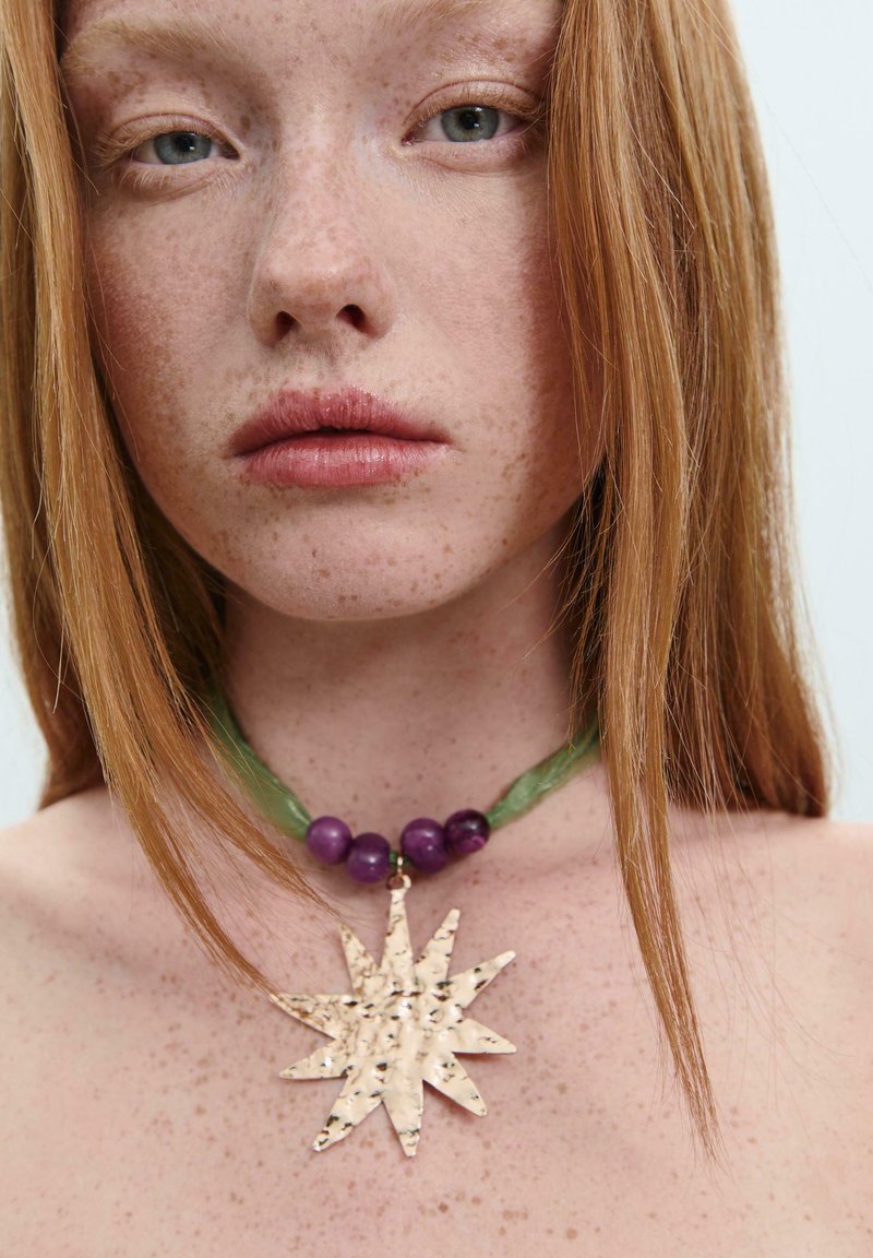 Ciondolo metallico a forma di stella su una collana con nastro verde e perle viola. Il ciondolo presenta una finitura testurizzata e martellata.