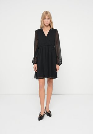Femme portant une robe noire à décolleté en V avec des manches longues transparentes et des escarpins slingback noirs à talon aigu, se tenant devant un fond blanc uni.