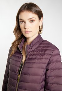 Jachetă pufoasă violetă cu închidere cu fermoar, având guler înalt, textură matlasată și accente de metal aurit.