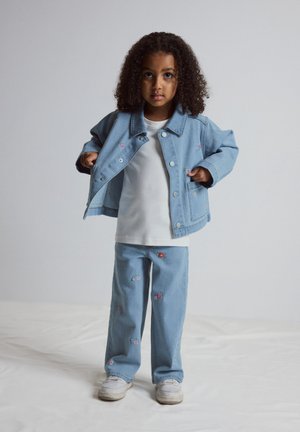 Kind met krullend haar dat een lichtblauwe spijkerjack en broek draagt met kleine bloemrijke borduursels, een wit T-shirt en witte sneakers.