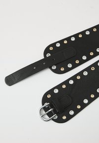 Black leather belt with a textured finish, featuring multiple metal eyelets in silver and gold, and a silver buckle detail.