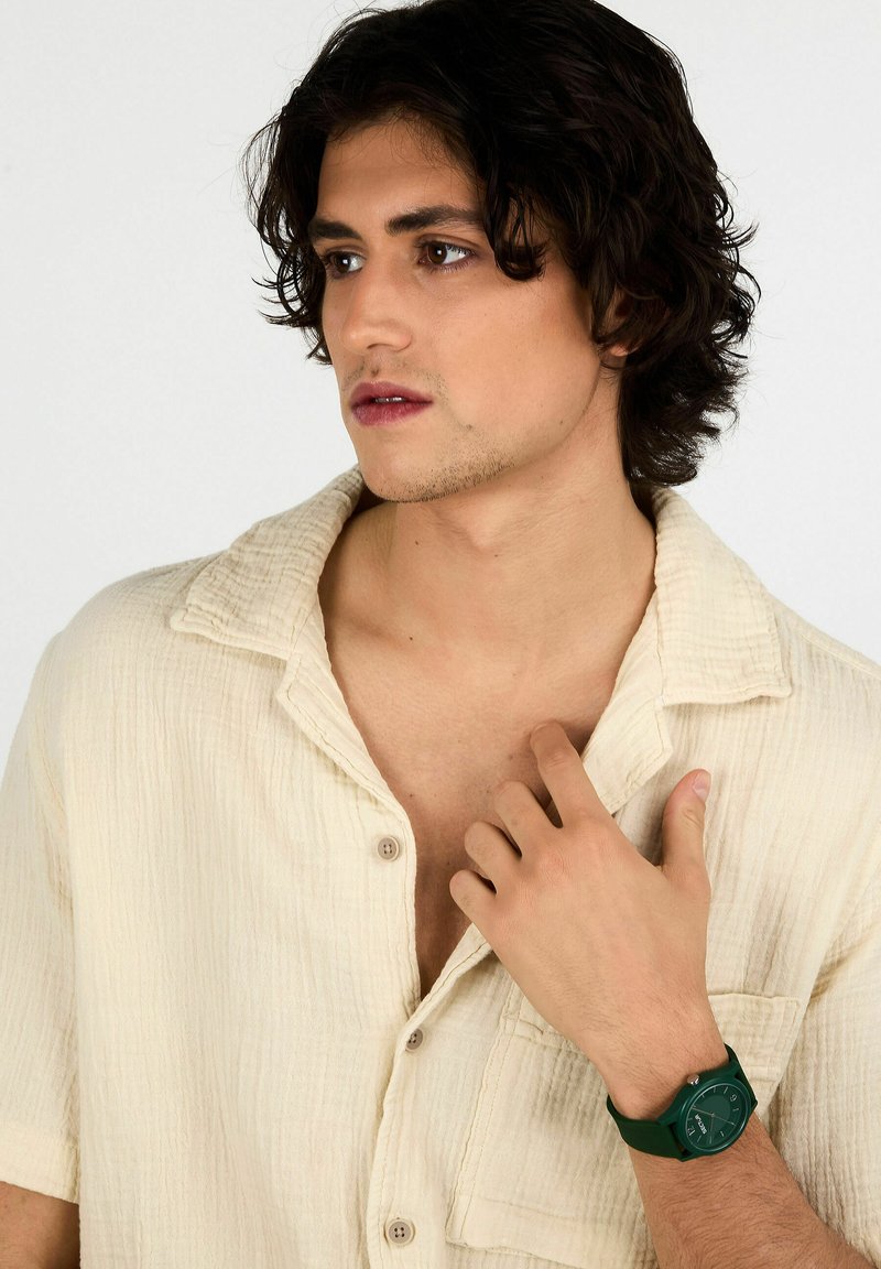 Orologio con cinturino in silicone verde indossato da un modello maschile, abbinato a una camicia a maniche corte in crema strutturata con colletto morbido e dettagli con bottoni.