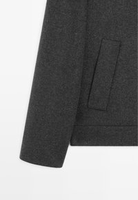 Dark gray wool jacket with a textured herringbone pattern. Features a single pocket and a tailored fit with a straight-cut sleeve.