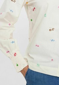 Blusa di leggero crema con ricami floreali colorati, polsini arricciati e orlo arricciato. Abbinata a jeans blu, che mostrano una vestibilità comoda.