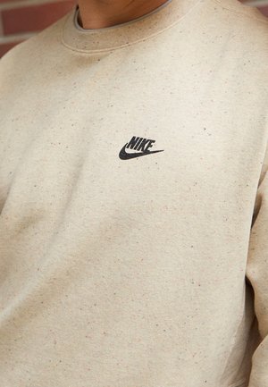 Beżowa melanżowa bluza z małym czarnym logo Nike na lewej piersi, noszona przez osobę na rozmytym tle.
