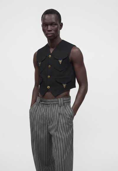 Homme mince à la peau foncée portant un gilet noir court sans manches avec des boutons dorés et un pantalon gris rayé taille haute, les mains dans les poches.